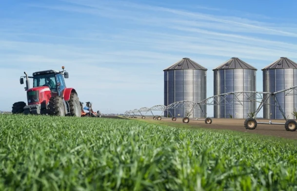 Trator vermelho em lavoura verde, com silos metálicos e pivô de irrigação ao fundo